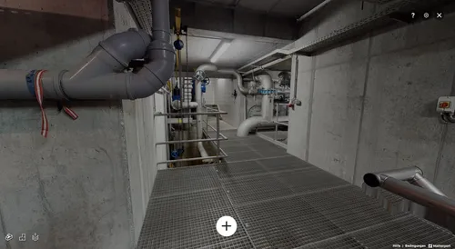 Ein Industriegebäude mit Rohren und Metallstrukturen, beleuchtet von einem hellen Deckenlicht. Eine Treppe führt in einen unteren Bereich.