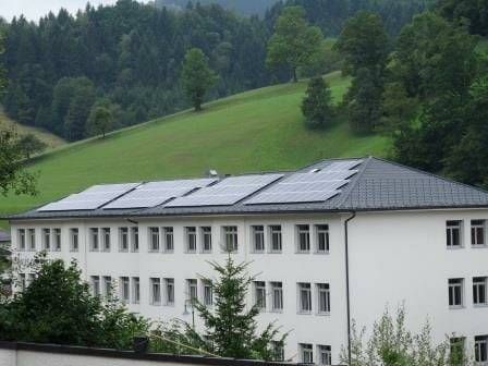 Ein weißes Gebäude mit Solarpanelen auf dem Dach und vielen Fenstern ist von grünen Bäumen und einem Hügel umgeben.