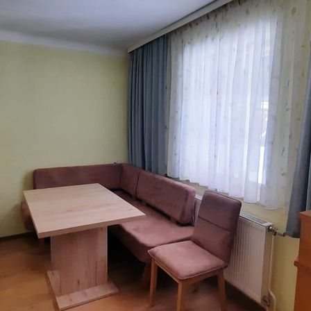 Eine gemütliche Ecke in einem Zimmer mit einem braunen Sofa, einem Holztisch und einem weißen Vorhang. Ein Heizkörper ist an der Wand montiert.
