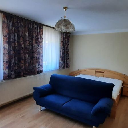 Ein Schlafzimmer mit einem blauen Sofa, einem Holzbett und einer Deckenleuchte. Der Raum hat eine weiße Wand und einen Holzboden.