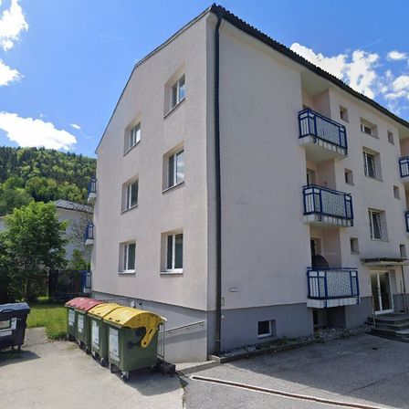 Ein mehrstöckiges Mehrfamilienhaus mit blauen Geländern und Fenstern, in einer Vorstadtgegend mit Rasenfläche und Mülleimern davor.