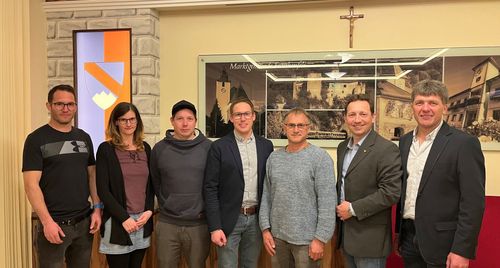 Fünf Personen stehen vor einer Wand mit einem Bild einer Kirche und einem Kreuz darüber. Sie alle lächeln und scheinen für ein Foto zu posieren.