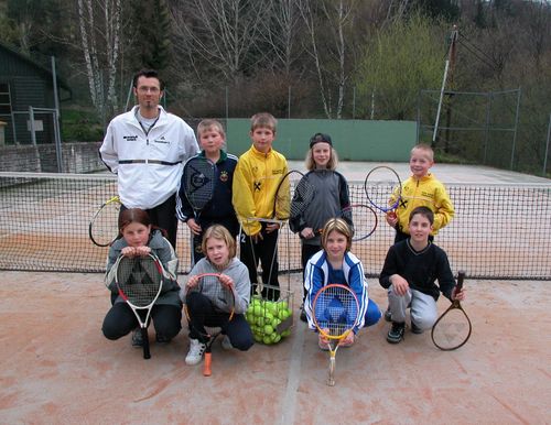 Eine Gruppe von Kindern und einem Mann posiert für ein Foto auf einem Tennisplatz mit Tennisschlägern und Tennisbällen im Vordergrund.