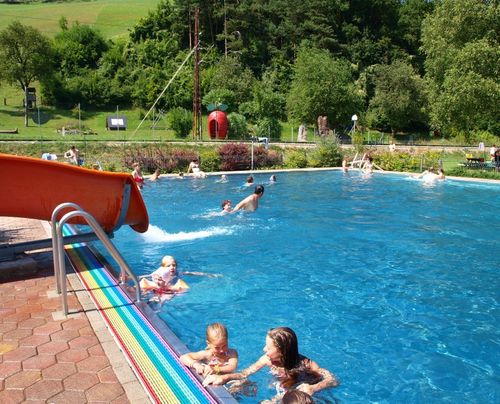 Eine Gruppe von Kindern und Erwachsenen schwimmt in einem großen Außenpool mit einer roten Rutsche und bunten Fliesen am Rand.