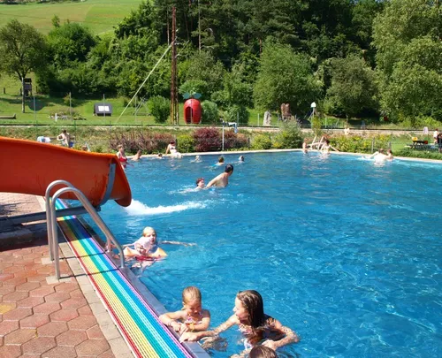 Eine Gruppe von Kindern und Erwachsenen schwimmt in einem großen Außenpool mit einer roten Rutsche und bunten Fliesen am Rand.
