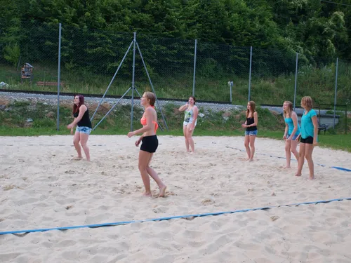Eine Gruppe junger Frauen spielt Beachvolleyball auf einem sandigen Spielfeld mit einem Netz, umgeben von einem Metallzaun und Grünpflanzen.