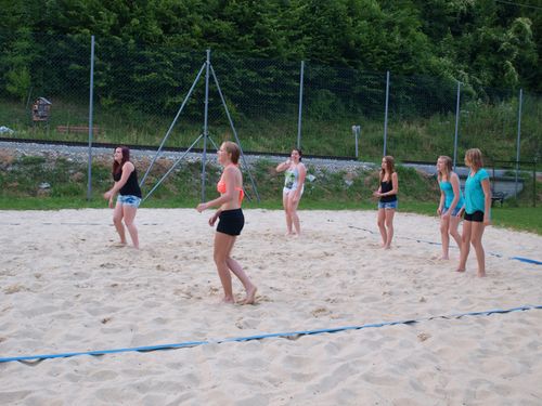 Eine Gruppe junger Frauen spielt Beachvolleyball auf einem sandigen Spielfeld mit einem Netz, umgeben von einem Metallzaun und Grünpflanzen.