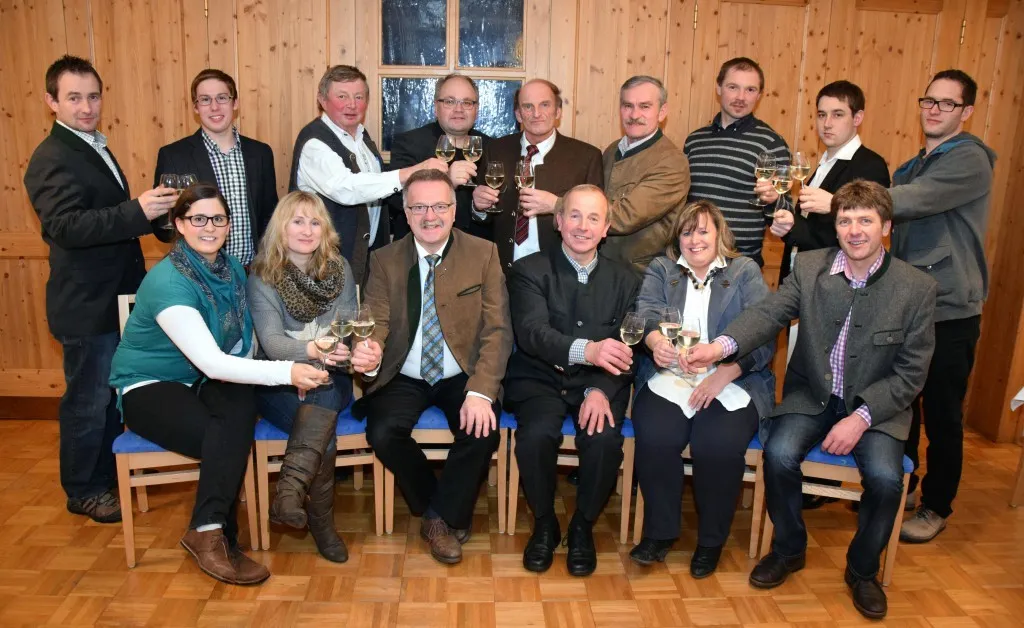 Eine Gruppe von Menschen, sowohl Männer als auch Frauen, sitzen auf Stühlen und halten Weingläser in der Hand, möglicherweise für ein Foto posend. Sie sind in formeller Kleidung gekleidet, und einige von ihnen tragen Brillen. Der Raum hat Holzwände und einen Holzboden.