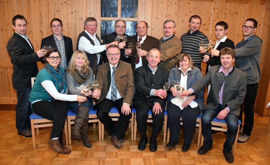 Eine Gruppe von Menschen, sowohl Männer als auch Frauen, sitzen auf Stühlen und halten Weingläser in der Hand, möglicherweise für ein Foto posend. Sie sind in formeller Kleidung gekleidet, und einige von ihnen tragen Brillen. Der Raum hat Holzwände und einen Holzboden.