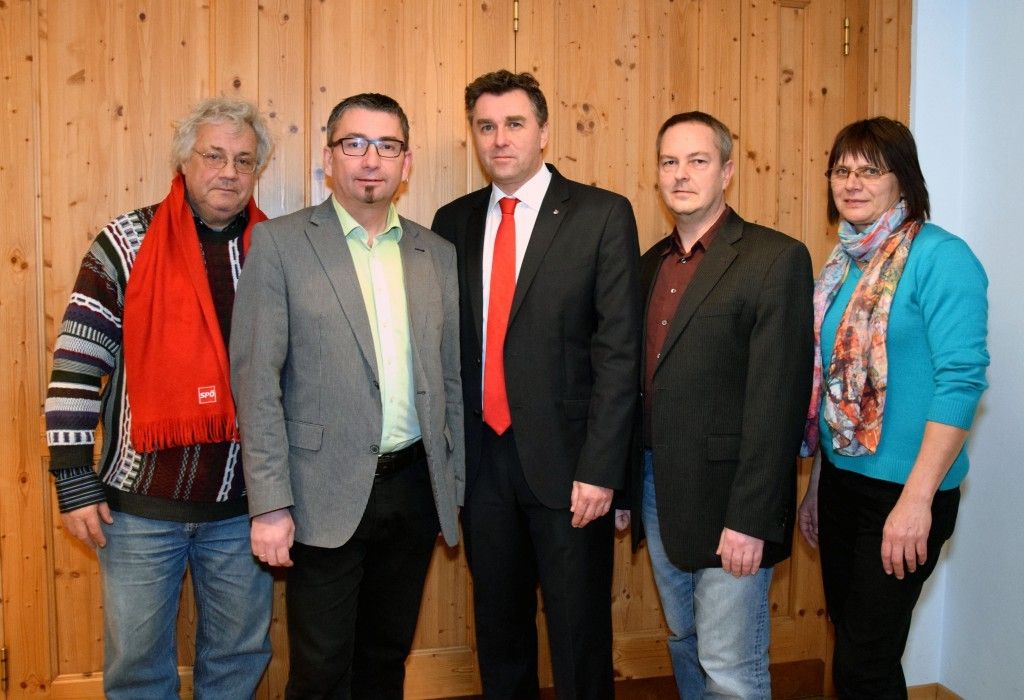 Fünf Personen, alle formell gekleidet, stehen zusammen vor einer Holzwand. Der Mann in der Mitte trägt eine rote Krawatte, während die anderen verschiedene Schattierungen von Schwarz und Grau tragen.