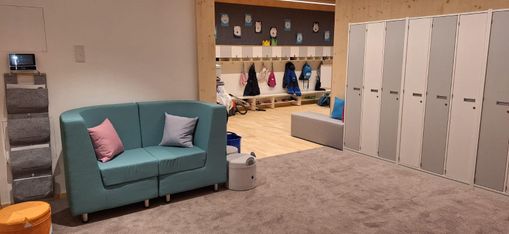 Ein Raum mit einer blauen Couch und einem grauen Kissen. Ein Teppichboden und eine Bank befinden sich rechts. Schließfächer und Regale an der Wand mit Taschen und Kleidung.