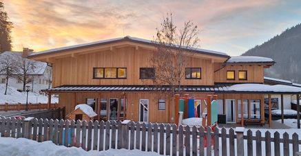 Ein Holzhaus mit schneebedecktem Dach, einem kleinen Hundehäuschen und einem Holzzaun davor. In der Nähe des Hauses befindet sich ein farbenfroher Spielplatz.