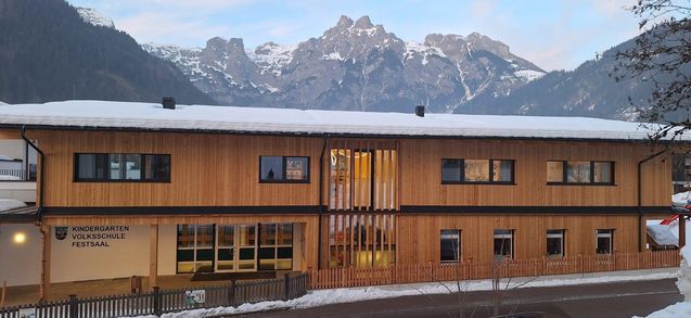Ein holzverkleidetes Gebäude mit Glasfenstern und Türen, umgeben von einem Holzzaun, unter einem verschneiten Bergrücken.