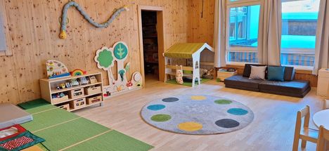 Ein Kinderspielzimmer mit Holzwänden, einem runden Teppich, einem Spielzeughaus, einem Bücherregal und Spielzeug. Eine Baum- und Pilzdekoration ist an der linken Wand.