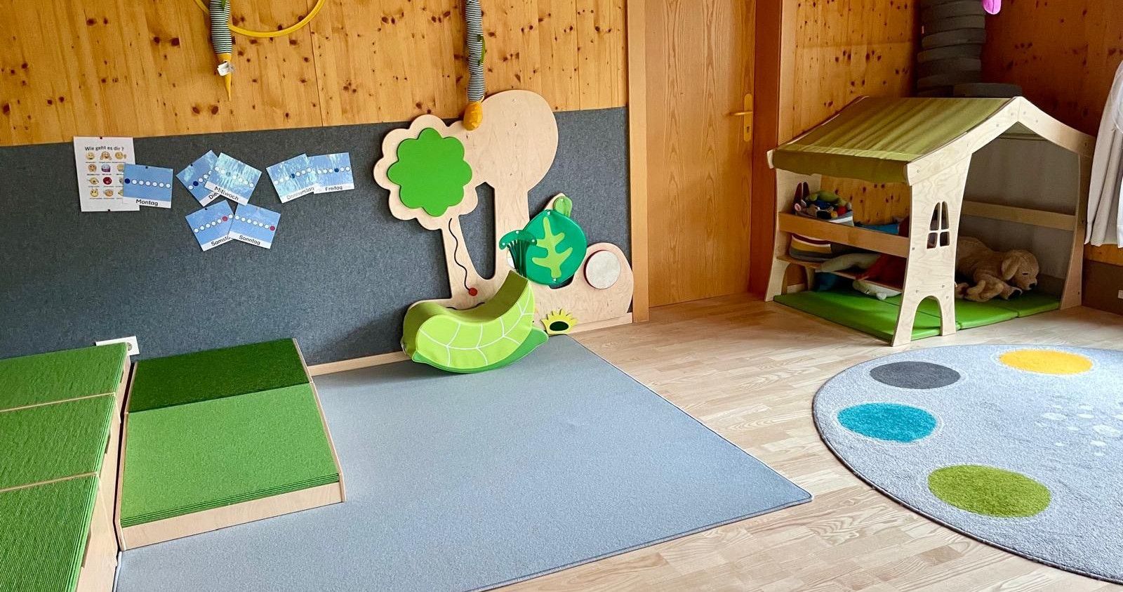 Ein Spielbereich für Kinder mit Holzwänden, einem grauen Teppich, einem Baum mit einer Schildkröte und einer Tür.