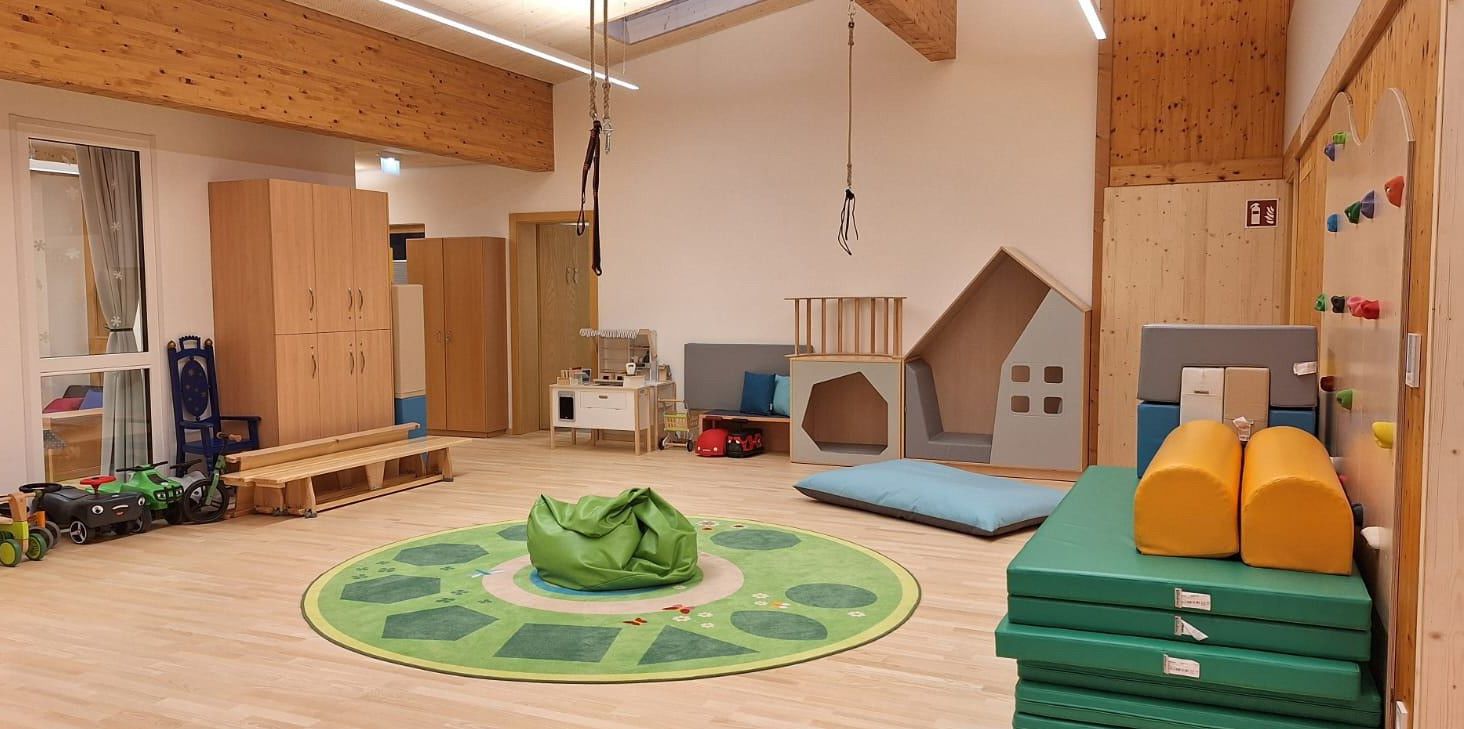 Ein Kinderspielzimmer mit Holzböden, mit einem grünen Sitzsack, rundem Teppich und verschiedenen Spielzeugen. Es gibt auch Schränke, einen kleinen Tisch und ein Puppenhaus. Zwei Schaukeln hängen von der Decke.