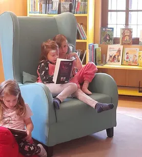 Drei junge Mädchen sitzen in einem blauen Stuhl in einer Bibliothek. Ein Mädchen liest ein Buch, ein anderes hält ein Tablet, und ein drittes Mädchen sitzt neben ihnen.