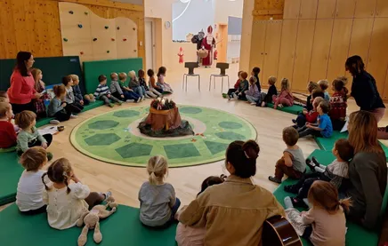 In einem Klassenzimmer sitzen Kinder in einem Kreis um einen grünen Teppich mit einer Weihnachtsmannfigur im Hintergrund. Ein Erwachsener sitzt mit einer Gitarre.