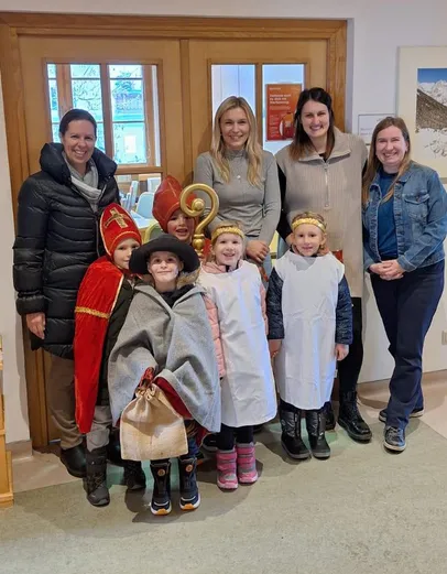Fünf Frauen und fünf Kinder in Kostümen posieren für ein Foto in einem Raum. Drei Kinder tragen Kronen, und die Erwachsenen lächeln. Ein Schild und ein Fenster sind im Hintergrund zu sehen.