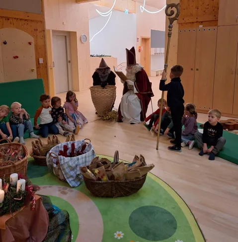 Ein Raum mit Kindern, die sich um den Nikolaus versammelt haben und Körbe mit Geschenken halten. Der Nikolaus liest aus einem Buch, und ein Elf steht neben ihm. Der Raum ist mit Holzwänden und einem grünen Teppich dekoriert.