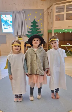 Drei Kinder in Kostümen stehen in einem Raum. Ein Mädchen links trägt ein weißes Kleid mit einem gelben Stirnband. Der Junge in der Mitte trägt einen grauen Mantel mit einem schwarzen Hut. Der Junge rechts trägt ein weißes Kleid mit einem goldenen Stirnband. Hinter ihnen steht ein Weihnachtsbaum und ein Fenster.