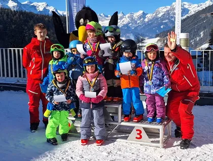 Eine Gruppe von Kindern steht auf einem Podest im Schnee, alle in Winterkleidung und mit Urkunden in der Hand. Dahinter sind ein Maskottchen und Erwachsene anwesend, mit verschneiten Bergen im Hintergrund.