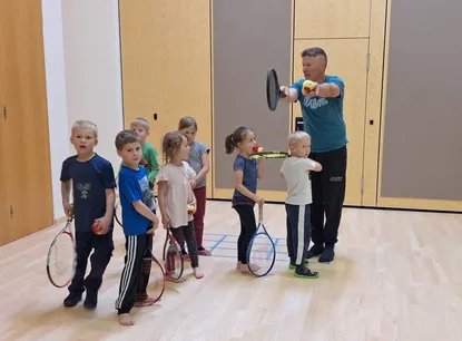 Eine Gruppe von Kindern lernt von einem Mann in einem blauen T-Shirt Tennis zu spielen. Sie halten Tennisschläger und Tennisbälle.