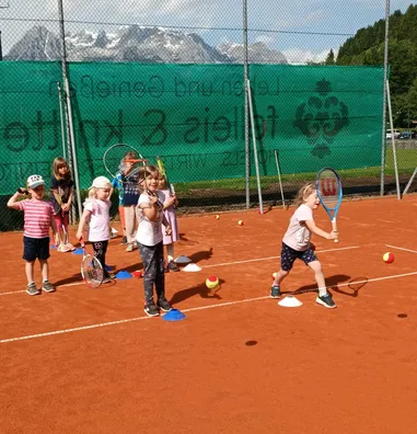 Eine Gruppe junger Kinder spielt Tennis auf einem Sandplatz mit Bergen im Hintergrund. Sie halten Schläger und stehen in der Nähe von Tennisbällen und Kegeln.