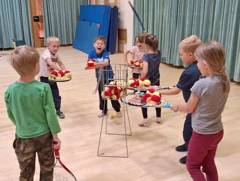 Kinder spielen in einer Turnhalle mit Tennisschlägern und schlagen Tennisbälle aus einem Korb. Sie tragen Freizeitkleidung und sind barfuß.