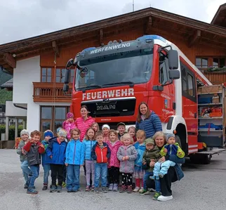 Eine Gruppe von Kindern und zwei Erwachsenen posiert lächelnd vor einem roten Feuerwehrauto. Dahinter steht ein Gebäude mit Holzbalkonen.