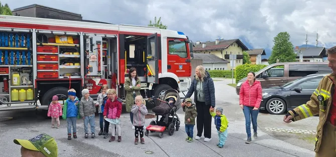 Eine Gruppe von Kindern und Erwachsenen steht vor einem Feuerwehrauto. Eine Frau hält ein Baby in einem Kinderwagen.