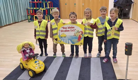 Eine Gruppe junger Kinder in Warnwesten steht auf einem schwarz-weißen Teppich und posiert für ein Foto mit einem Schild, das 'Das kleine Straßen 1x1' sagt.