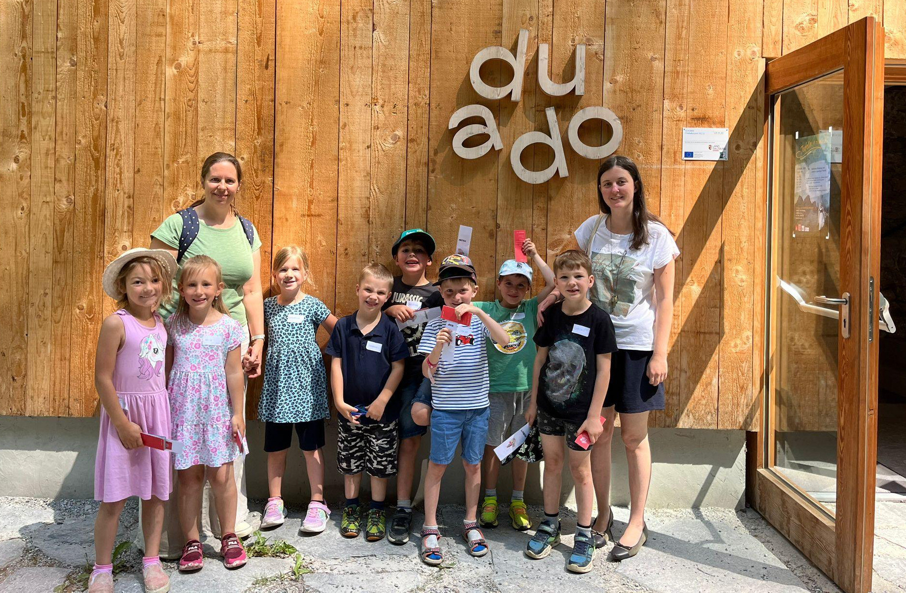 Eine Gruppe von Kindern und zwei Frauen steht vor einer Holzwand mit dem Wort 'dudo' darauf. Sie haben alle Namensschilder.