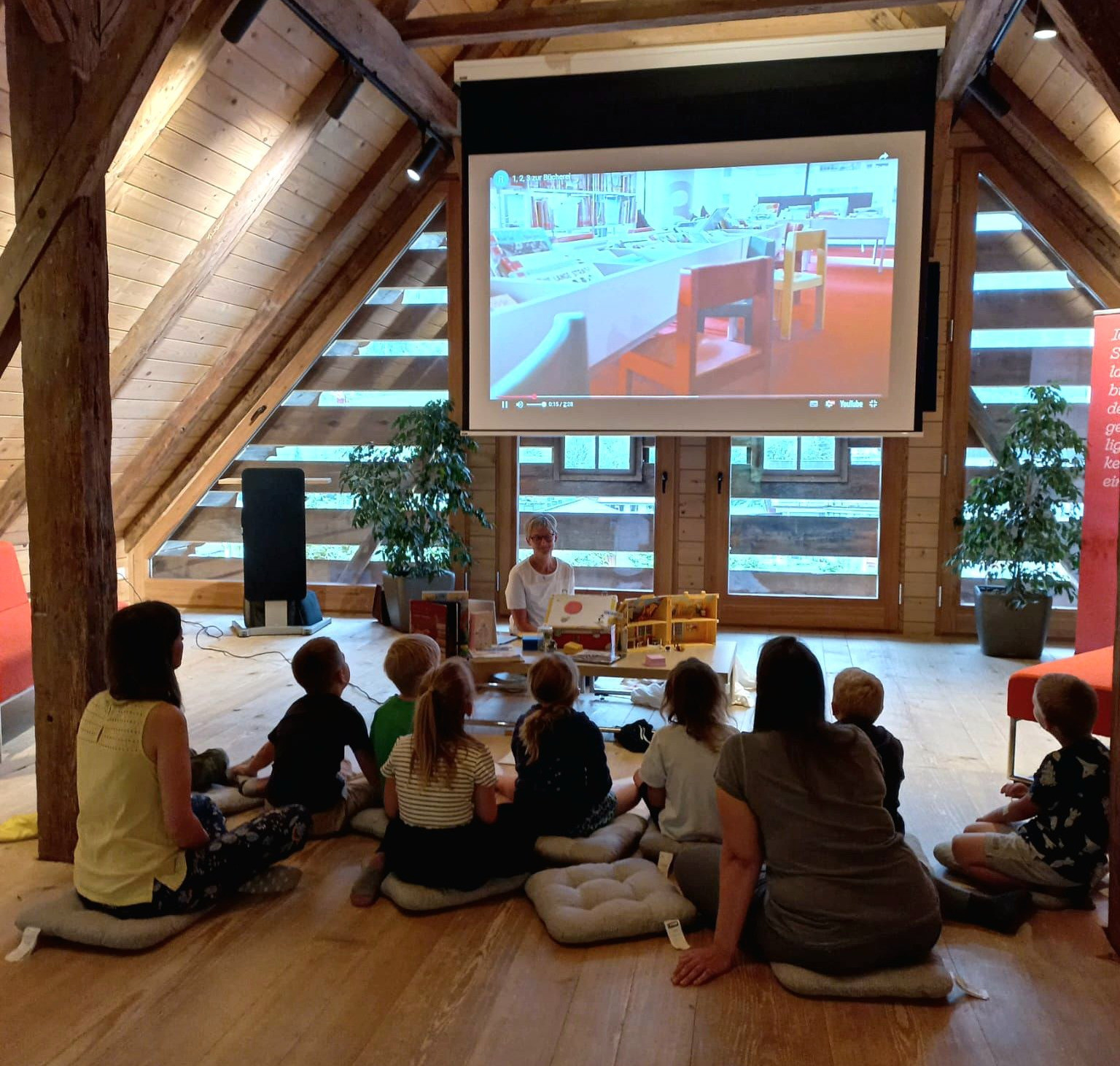 In einem Dachzimmer hält eine Frau eine Präsentation für eine Gruppe von Kindern ab. Die Projektionsleinwand zeigt ein Bibliotheksinneres. Die Kinder sitzen auf Kissen und hören aufmerksam zu.