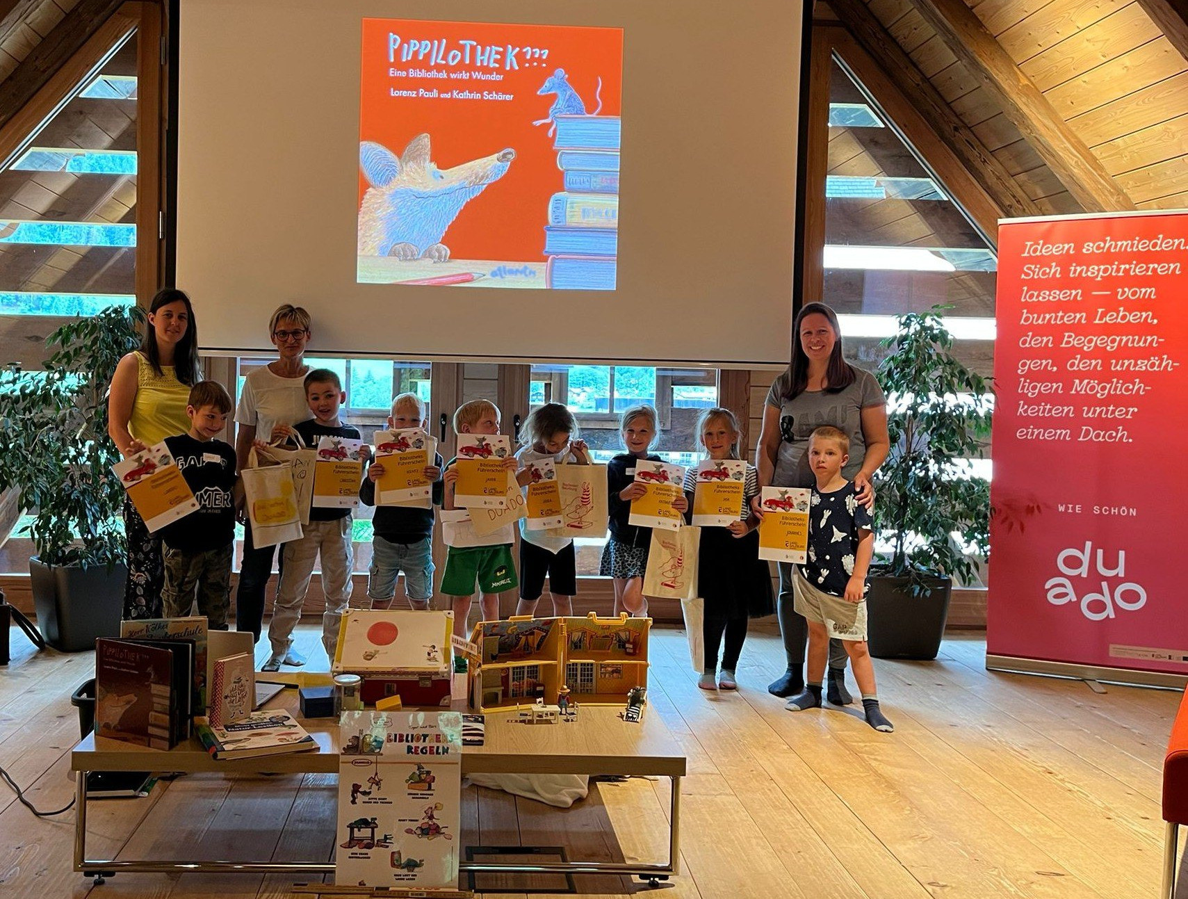 Kinder halten Urkunden in der Hand, während sie in einem Raum mit Beamerleinwand, Büchern und einer lächelnden Frau stehen.