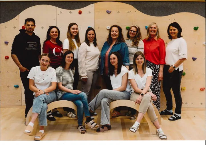 Eine Gruppe von Frauen und Männern posiert für ein Foto in einem Fitnessstudio. Sie sitzen auf hölzernen Bänken und hinter ihnen steht eine Kletterwand.