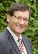 Ein Mann in Anzug und Krawatte, mit Brille, lächelt fröhlich für ein Porträt vor einem unscharfen Hintergrund aus grünem Laub.