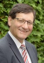 Ein Mann in Anzug und Krawatte, mit Brille, lächelt fröhlich für ein Porträt vor einem unscharfen Hintergrund aus grünem Laub.