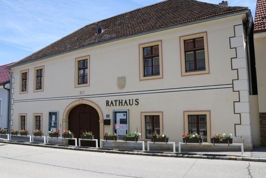 Ein traditionelles europäisches Gebäude mit dem Wort Rathaus auf der Vorderseite, mit einem großen Bogeneingang, mehreren Fenstern und Blumenkästen darunter.