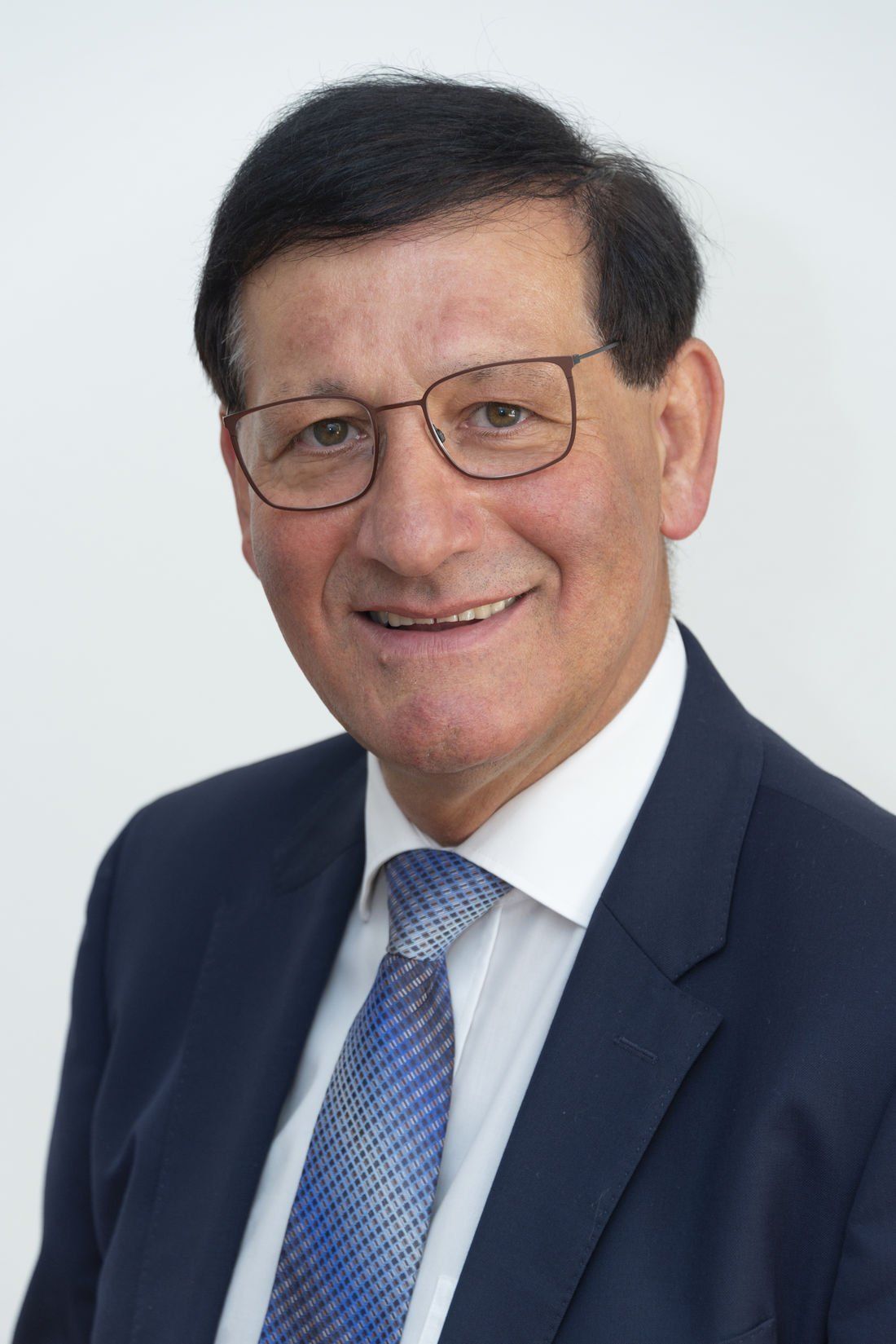 Ein Mann in Anzug und Krawatte, mit Brille, lächelt in die Kamera. Er hat kurzes, dunkles Haar und ist glattrasiert.