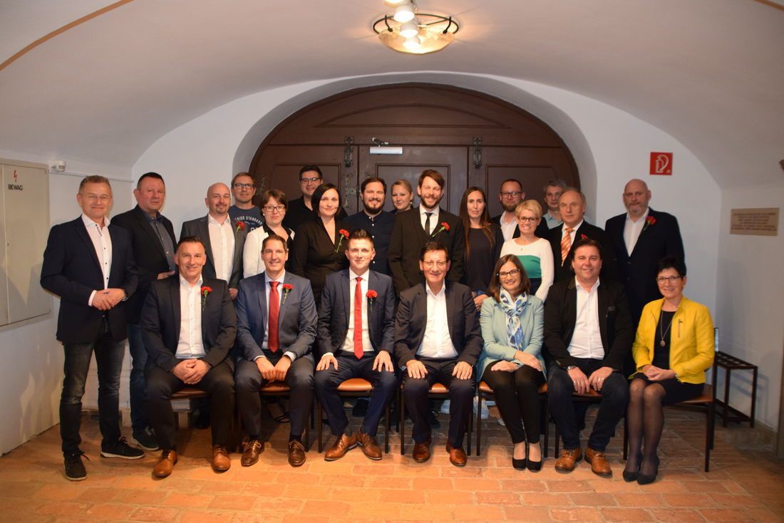 Eine Gruppe formell gekleideter Personen, einige sitzend und andere stehend, posiert für ein Foto in einem Raum mit Ziegelboden und einem hölzernen Bogen.