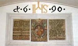 Ein gerahmtes Schild mit goldenem Text 'd.6.1990' über drei Tafeln mit altem Text, eine zentrale mit einem Bild eines gekrönten Mannes.