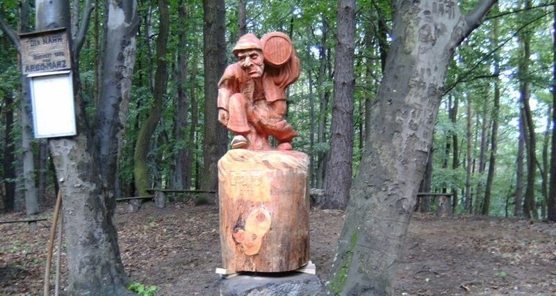 Eine Holzstatue eines Mannes, der auf einem Baumstumpf in einem Wald sitzt und einen Eimer hält.