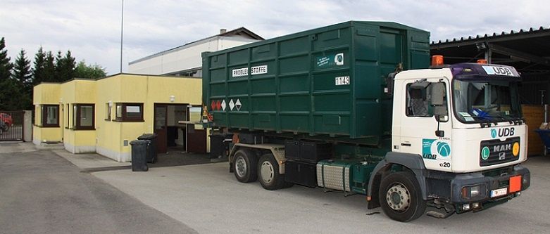 Ein großer grüner und weißer LKW ist vor einem gelben Gebäude mit mehreren Fenstern und einem Tor geparkt. Der LKW hat einen großen Container angebaut.