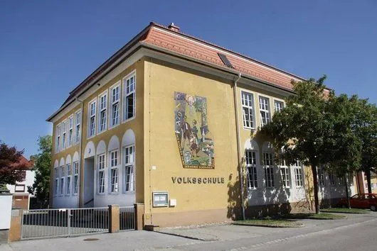 Ein gelbes Gebäude mit weißen Fensterrahmen und mehreren Fenstern, beschriftet mit 'Volksschule', steht an einer Straße, wo ein rotes Auto geparkt ist.