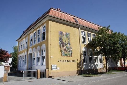 Ein gelbes Gebäude mit weißen Fensterrahmen und mehreren Fenstern, beschriftet mit 'Volksschule', steht an einer Straße, wo ein rotes Auto geparkt ist.