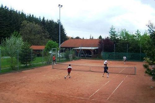 Vier Personen spielen Tennis auf einem Sandplatz, der durch ein Netz in zwei Hälften geteilt ist. Bäume und ein Gebäude sind im Hintergrund zu sehen.