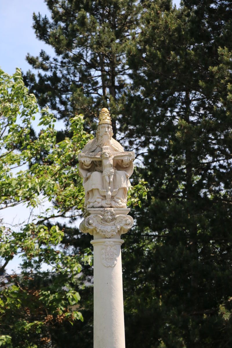 Eine Steinstatue einer gekrönten Figur, die ein Buch und ein Kind hält, steht auf einer Säule mit verzierten Schnitzereien. Die Statue steht vor einem Hintergrund aus grünen Bäumen.