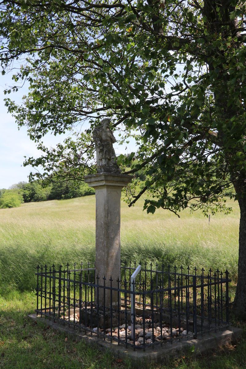 Ein verwittertes Steinmonument mit einer Statue einer geflügelten Figur steht auf einer Wiese, umgeben von einem schwarzen Eisenzaun unter einem blattreichen Baum.