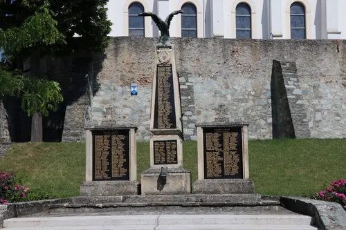 Ein Denkmal mit drei Steinsäulen, die jeweils mit Namen graviert sind, steht vor einer Steinmauer mit Bogenfenstern. Auf der mittleren Säule thront eine Adlerstatue.